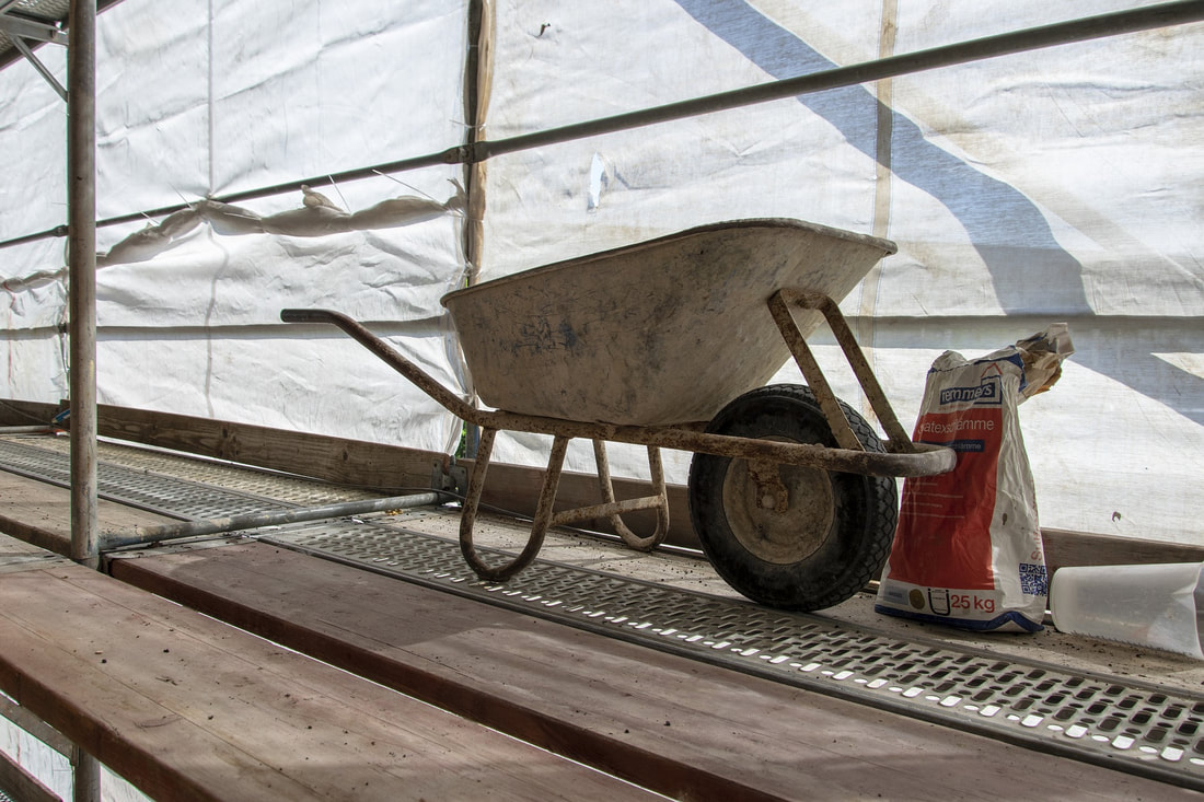 Picture of a wheel barrow on a scaffold platform. Its day time and the sunlight is casting shadows on the platform through a non-see through white plastic sheet brick guard. There is a bag of dry construction cement next to the wheel barrow.