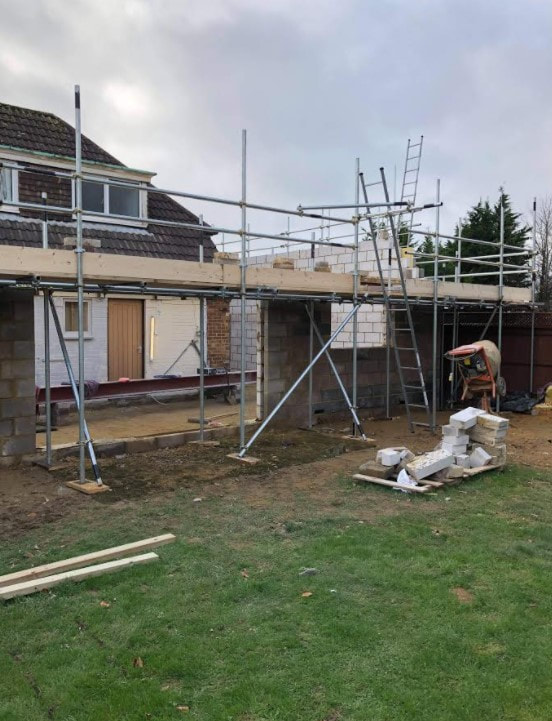  A picture of the rear of a bungalow that has been taken from the garden. An extension is being built on the back of the property in blockwork. The extension ha been built up to first floor level and there is a scaffold up to the top of the extension build so far.