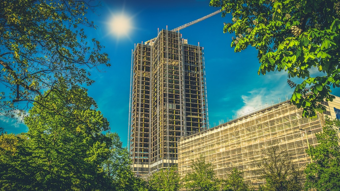 Picture of a high rise tower block and multi storey tower block. Both buildings have scaffolding around the outside and are undergoing construction work. It is a bright sunny day, with clear blue skies