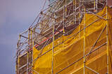 Picture a commercial scaffold on a high rise building with a yellow mesh brick guard. It is a sunny day with blue sky.