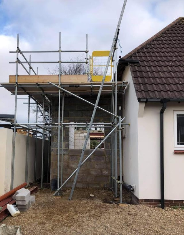 Picture of the side of a white rendered house that is having an extension built. The extension is mid-build and there is scaffolding up to the top of the first storey. 