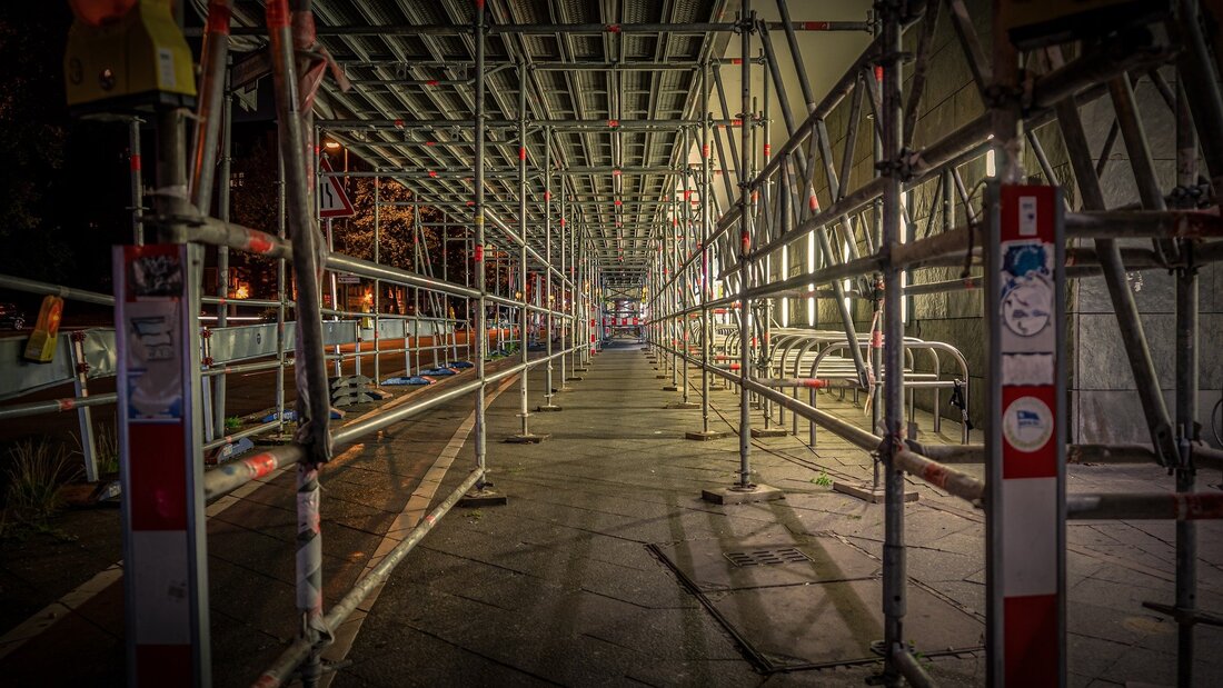 Picture a gantry scaffold over a public street. There is a walkway lined either side with scaffolding and guard rails. It is night time and the walkway is well lit with artificial lighting